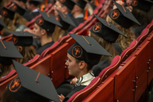 A group of people in black academic gowns and mortarboards with orange symbols are seated in rows in the auditorium during an academic ceremony.
