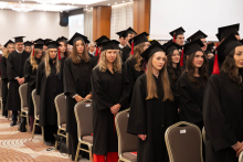People in black academic gowns and mortarboards stand beside chairs in the hall during an academic ceremony.