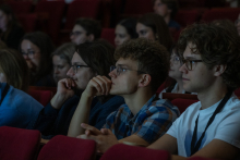 Participants sit in rows in the auditorium and listen to the presentation. In the foreground, three people are focused on the presentation, while more attendees are seated in the background.