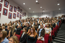 A large group of ceremony participants stand among rows of red seats in an auditorium. A gallery of framed black-and-white portraits is displayed on the wall to the left.