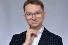 Portrait of a person wearing glasses, a white shirt and a dark patterned jacket, against a light grey background.