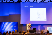 Mężczyzna stoi przy mównicy i prowadzi wystąpienie. Na sali siedzą uczestnicy konferencji. Obok sceny znajdują się plansze informacyjne organizatora i partnerów wydarzenia.
