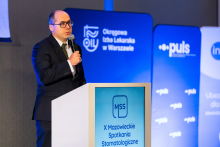 Mężczyzna - prezes OIL przemawia przy mównicy podczas X Mazowieckich Spotkań Stomatologicznych. Przed sceną siedzą uczestnicy wydarzenia. W tle widoczne są plansze z logo Okręgowej Izby Lekarskiej w Warszawie oraz partnerów wydarzenia.
