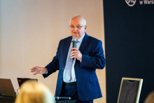 Mężczyzna prowadzi wykład. W ręku trzyma mikrofon, a drugą dłoń podnosi lekko do góry. Za nim widać kawałek ścianki organizatora, przed nim - słuchaczy.