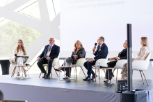 Sześć osób uczestniczy w panelu dyskusyjnym na scenie. Paneliści siedzą w fotelach i korzystają z mikrofonów. Przed nimi ustawiono stoliki z wodą.