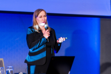 Kobieta prowadzi wykład ze sceny. Za nią wyświetlana jest prezentacja, a przed sceną siedzą uczestnicy konferencji.