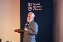 Mężczyzna prowadzi wykład. Mówi do mikrofonu. Za nim widać ściankę organizatora wydarzenia.