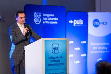 Mężczyzna prowadzi wykład na scenie. Stoi przy mównicy i przemawia do mikrofonu.