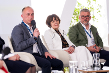 Trzy osoby uczestniczą w panelu dyskusyjnym. Jedna osoba mówi do mikrofonu, a pozostałe siedzą obok na scenie.