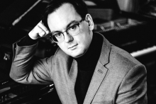 Black-and-white portrait of a person wearing glasses and a jacket, seated beside a piano with their head resting on one hand.