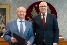 Dwóch elegancko ubranych mężczyzn pozuje do zdjęcia. Uśmiechają się do zdjęcia. Mężczyzna po lewej trzyma zamkniętą teczkę z dokumentem. W tle znajduje się sala posiedzeń z uczestnikami wydarzenia.