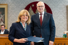 Elegancko ubrani kobieta i mężczyzna pozują do zdjęcia. Uśmiechają się do zdjęcia. Kobieta trzyma zamkniętą teczkę z dokumentem. W tle znajduje się sala posiedzeń z uczestnikami wydarzenia.