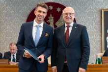 Dwóch elegancko ubranych mężczyzn pozuje do zdjęcia. Uśmiechają się do zdjęcia. Mężczyzna po lewej trzyma zamkniętą teczkę z dokumentem. W tle znajduje się sala posiedzeń z uczestnikami wydarzenia.