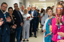 Kilkanaście osób stoi po obu stronach szpitalnego korytarza. Część uczestników klaszcze. W centrum widoczne są osoby w stroju medycznym i formalnym podczas oficjalnego wydarzenia.