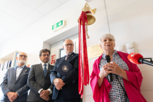 Cztery osoby stoją przy ścianie obok mosiężnego dzwonu z czerwonymi wstążkami. Jedna osoba przemawia do mikrofonu. Pod dzwonem znajduje się tabliczka pamiątkowa.