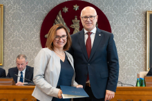 Elegancko ubrani kobieta i mężczyzna pozują do zdjęcia. Uśmiechają się do zdjęcia. Kobieta trzyma zamkniętą teczkę z dokumentem. W tle znajduje się sala posiedzeń z uczestnikami wydarzenia.