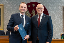 Dwóch elegancko ubranych mężczyzn pozuje do zdjęcia. Uśmiechają się do zdjęcia. Mężczyzna po lewej trzyma zamkniętą teczkę z dokumentem. W tle znajduje się sala posiedzeń z uczestnikami wydarzenia.