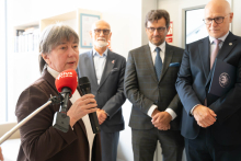Jedna osoba przemawia do mikrofonu. W tle stoją trzy osoby uczestniczące w wydarzeniu. Widoczny jest fragment dzwonu i wyposażenia pomieszczenia.