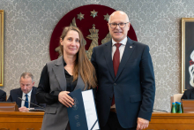 Elegancko ubrani kobieta i mężczyzna pozują do zdjęcia. Uśmiechają się do zdjęcia. Kobieta trzyma otwartą teczkę z dokumentem. W tle znajduje się sala posiedzeń z uczestnikami wydarzenia.