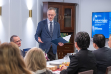 Dziekan przemawia podczas spotkania stojąc za stołem konferencyjnym. W ręce trzyma otwartą teczkę. Wokół siedzą pozostali uczestnicy.