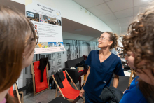 Uczestnicy Targów Kół Naukowych rozmawiają przy stoisku Studenckiego Koła Naukowego Terapii Płodu i Perinatologii. W tle znajduje się plansza prezentująca działalność koła.