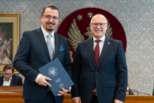 Dwóch elegancko ubranych mężczyzn pozuje do zdjęcia. Uśmiechają się do zdjęcia. Mężczyzna po lewej trzyma zamkniętą teczkę z dokumentem. W tle znajduje się sala posiedzeń z uczestnikami wydarzenia.
