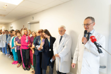 Grupa kilkudziesięciu osób stoi w szpitalnym korytarzu. Jedna osoba w białym fartuchu przemawia do mikrofonu, a pozostali uczestnicy słuchają wystąpienia.
