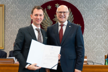 Dwóch elegancko ubranych mężczyzn pozuje do zdjęcia. Uśmiechają się do zdjęcia. Mężczyzna po lewej trzyma otwartą teczkę z dokumentem. W tle znajduje się sala posiedzeń z uczestnikami wydarzenia.