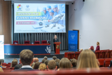 Kobieta przemawia przy mównicy w sali audytoryjnej. Uczestnicy siedzą na widowni i słuchają wystąpienia. Na dużym ekranie wyświetlona jest informacja o I Mistrzostwach Polski w Chirurgii Małoinwazyjnej.
