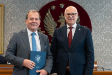 Dwóch elegancko ubranych mężczyzn pozuje do zdjęcia. Uśmiechają się do zdjęcia. Mężczyzna po lewej trzyma zamkniętą teczkę z dokumentem. W tle znajduje się sala posiedzeń z uczestnikami wydarzenia.