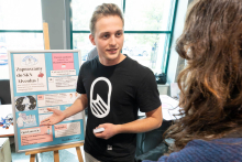 Osoba reprezentująca Studenckie Koło Naukowe Alveolus rozmawia z uczestnikiem Targów Kół Naukowych. W tle znajduje się plansza informująca o działalności koła.
