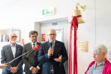 Trzy osoby stoją przy ścianie obok Dzwonu Życia. Jedna osoba przemawia do mikrofonu. Widoczna jest tabliczka informacyjna umieszczona pod dzwonem.