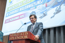 Mężczyzna stoi przy mównicy i przemawia podczas wydarzenia na sali audytoryjnej. Za mównicą widoczny jest ekran z informacją o I Mistrzostwach Polski w Chirurgii Małoinwazyjnej organizowanych 16-17 października 2026 r.