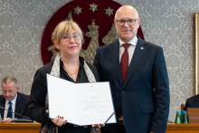 Elegancko ubrani kobieta i mężczyzna pozują do zdjęcia. Uśmiechają się do zdjęcia. Kobieta trzyma otwartą teczkę z dokumentem. W tle znajduje się sala posiedzeń z uczestnikami wydarzenia.