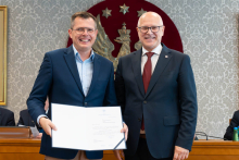 Dwóch elegancko ubranych mężczyzn pozuje do zdjęcia. Uśmiechają się do zdjęcia. Mężczyzna po lewej trzyma otwartą teczkę z dokumentem. W tle znajduje się sala posiedzeń z uczestnikami wydarzenia.