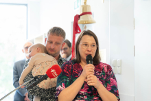 Dwie osoby stoją przy mikrofonach podczas wydarzenia. Jedna osoba przemawia, druga trzyma na rękach małe dziecko. W tle widoczny jest dzwon zamontowany na ścianie.