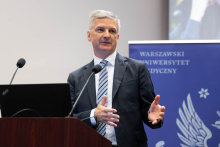Mężczyzna przy mównicy z laptopem i mikrofonami. W tle widoczny jest ekran prezentacji z napisem „Everything is Chemistry” oraz portretem prelegenta wydarzenia.