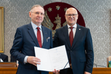Dwóch elegancko ubranych mężczyzn pozuje do zdjęcia. Uśmiechają się do zdjęcia. Mężczyzna po lewej trzyma otwartą teczkę z dokumentem. W tle znajduje się sala posiedzeń z uczestnikami wydarzenia.