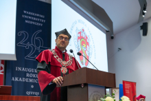 Dziekan w stroju akademickim przemawia przy mównicy. W tle widoczny jest ekran z herbem uczelni oraz baner inauguracji roku akademickiego.