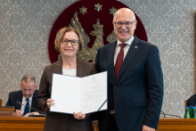 Elegancko ubrani kobieta i mężczyzna pozują do zdjęcia. Uśmiechają się do zdjęcia. Kobieta trzyma otwartą teczkę z dokumentem. W tle znajduje się sala posiedzeń z uczestnikami wydarzenia.