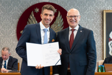 Dwóch elegancko ubranych mężczyzn pozuje do zdjęcia. Uśmiechają się do zdjęcia. Mężczyzna po lewej trzyma otwartą teczkę z dokumentem. W tle znajduje się sala posiedzeń z uczestnikami wydarzenia.