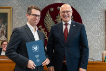 Dwóch elegancko ubranych mężczyzn pozuje do zdjęcia. Uśmiechają się do zdjęcia. Mężczyzna po lewej trzyma zamkniętą teczkę z dokumentem. W tle znajduje się sala posiedzeń z uczestnikami wydarzenia.