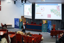 Noblista stoi przed publicznością w auli i wskazuje ręką na prezentację. Na ekranie widnieje slajd „Chemistry is everything… Life is complex but logically organized” z wzorami chemicznymi i modelami cząsteczek. Obok sceny stoją materiały Warszawskiego Uniwersytetu Medycznego i wykładu „Everything is Chemistry”.