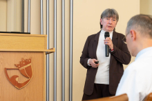Jedna osoba wygłasza prezentację przy mównicy, trzymając mikrofon i pilot do zmiany slajdów.