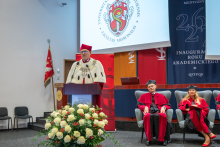 Rektor w stroju akademickim przemawia przy mównicy. Za nim znajdują się przedstawiciele władz dziekańskich siedzący na scenie oraz ekran z herbem wydziału.