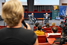 Jedna osoba na pierwszym planie trzyma mikrofon. Przed publicznością dwie osoby siedzą w fotelach na scenie. Obok stoi baner wydarzenia „Everything is Chemistry” oraz baner Warszawskiego Uniwersytetu Medycznego.