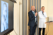 Dwie osoby stoją obok mównicy. Jedna przemawia do mikrofonu, druga słucha wystąpienia.