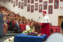 Rektor w stroju akademickim przemawia do uczestników zgromadzonych w auli. W tle widoczne są rzędy siedzeń zajęte przez publiczność.