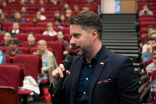 Mężczyzna stojący wśród publiczności zadaje pytanie przez mikrofon. W tle uczestnicy siedzą w rzędach czerwonych foteli auli.