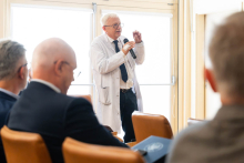 Jedna osoba stojąca między rzędami krzeseł zabiera głos podczas dyskusji. W pierwszym planie widoczni są uczestnicy spotkania. Przekaż swoją opinię na temat programu BizChat